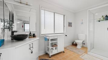 A bathroom with a white sink, mirror, and toilet. There is a window with blinds, a shower area, and a small shelf with toiletries.