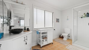 A bathroom with a white sink, mirror, and toilet. There is a window with blinds, a shower area, and a small shelf with toiletries. - Panorama Bay Escape San Remo (San Remo)