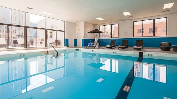 Una piscina interior moderna con agua azul cristalina, ventanas amplias y sillones de descanso.