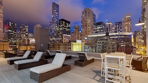 Un salón en la azotea con sillones y una mesa, con vistas a un horizonte urbano al atardecer.
