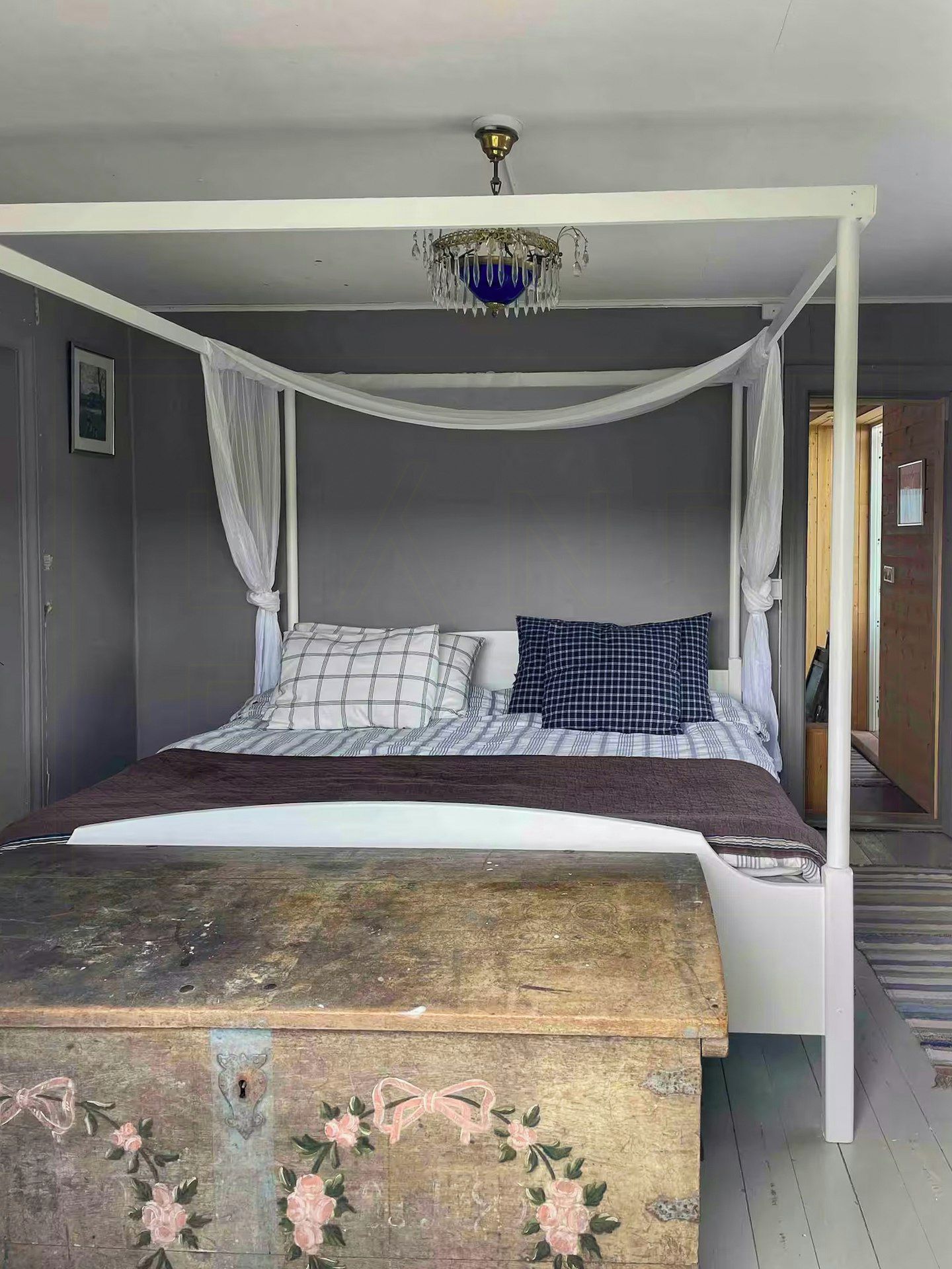 A four-poster bed with a canopy, a wooden chest with floral artwork, and a ceiling light fixture.