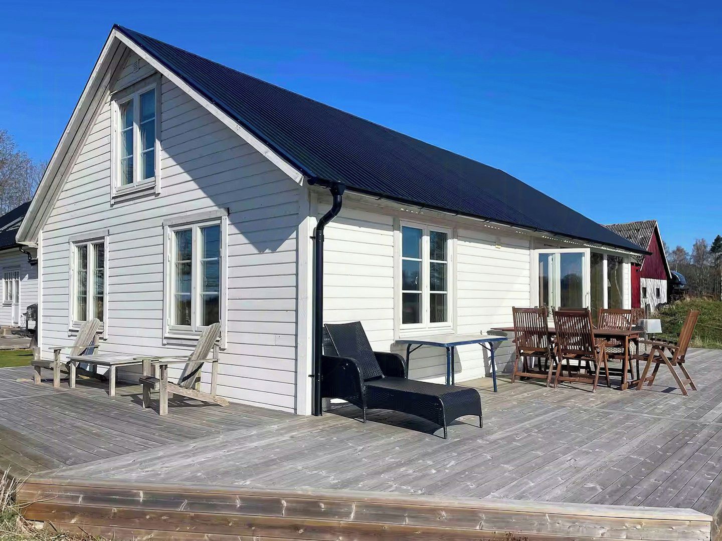 Een wit houten huis met een donker dak, een terras met tuinmeubilair en een rode schuur op de achtergrond.