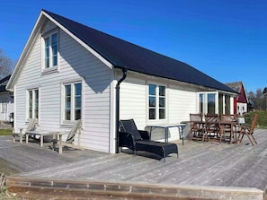 A white wooden house with a dark roof, a deck with outdoor furniture, and a red barn in the background.