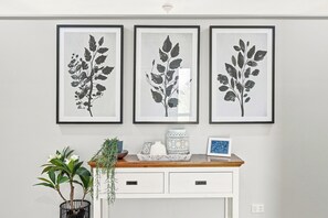 A wall with three framed botanical prints, a wooden console table with decorative items, and a potted plant. - HiTide at Toogoom | Stunning Views | Air-Con | Beach Access | Linens | Pets (Toogoom)