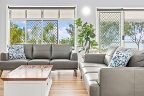 Ein Wohnzimmer mit grauen Sofas, einem Holztisch und großen Fenstern mit Blick auf Grünflächen und ein Gewässer.