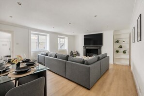 A modern living room with a grey sectional sofa, a flat-screen TV mounted above a fireplace, and a dining area with a glass table and chairs. - 2-bed Flat Near Regent s Park - Patio - Sleeps 6 (London)