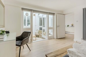 A modern living room with a glass desk, a black chair, and a white sofa. There is a sliding glass door leading to a balcony with outdoor furniture. - 2-bed Flat Near Regent s Park - Patio - Sleeps 6 (London)