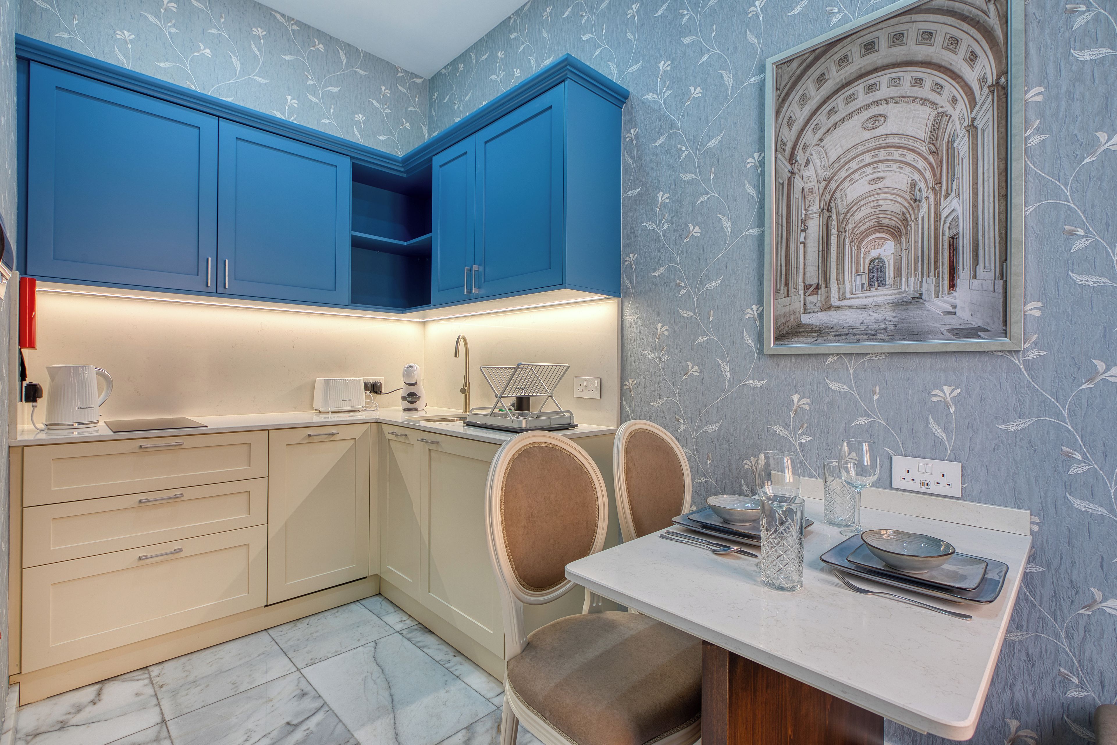 Une cuisine moderne avec des meubles bleus, une table à manger avec des chaises et un mur décoratif orné d’un tableau encadré.