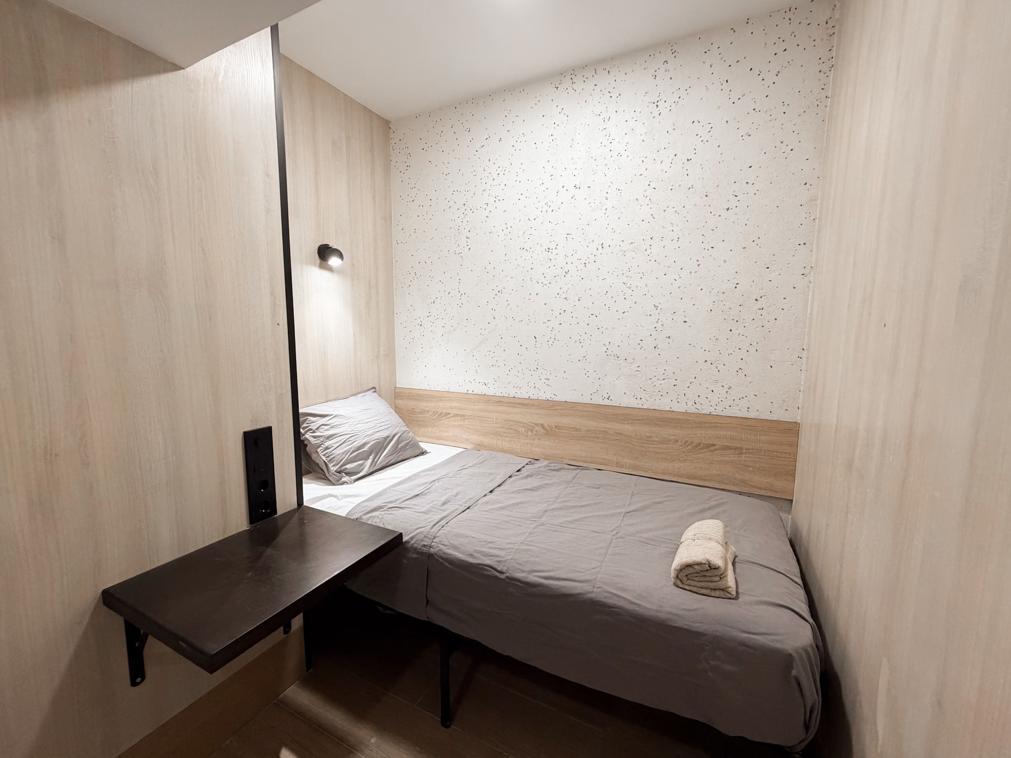 Pequeña habitación con cama individual, cabecera de madera, mesita de noche y luz de pared.