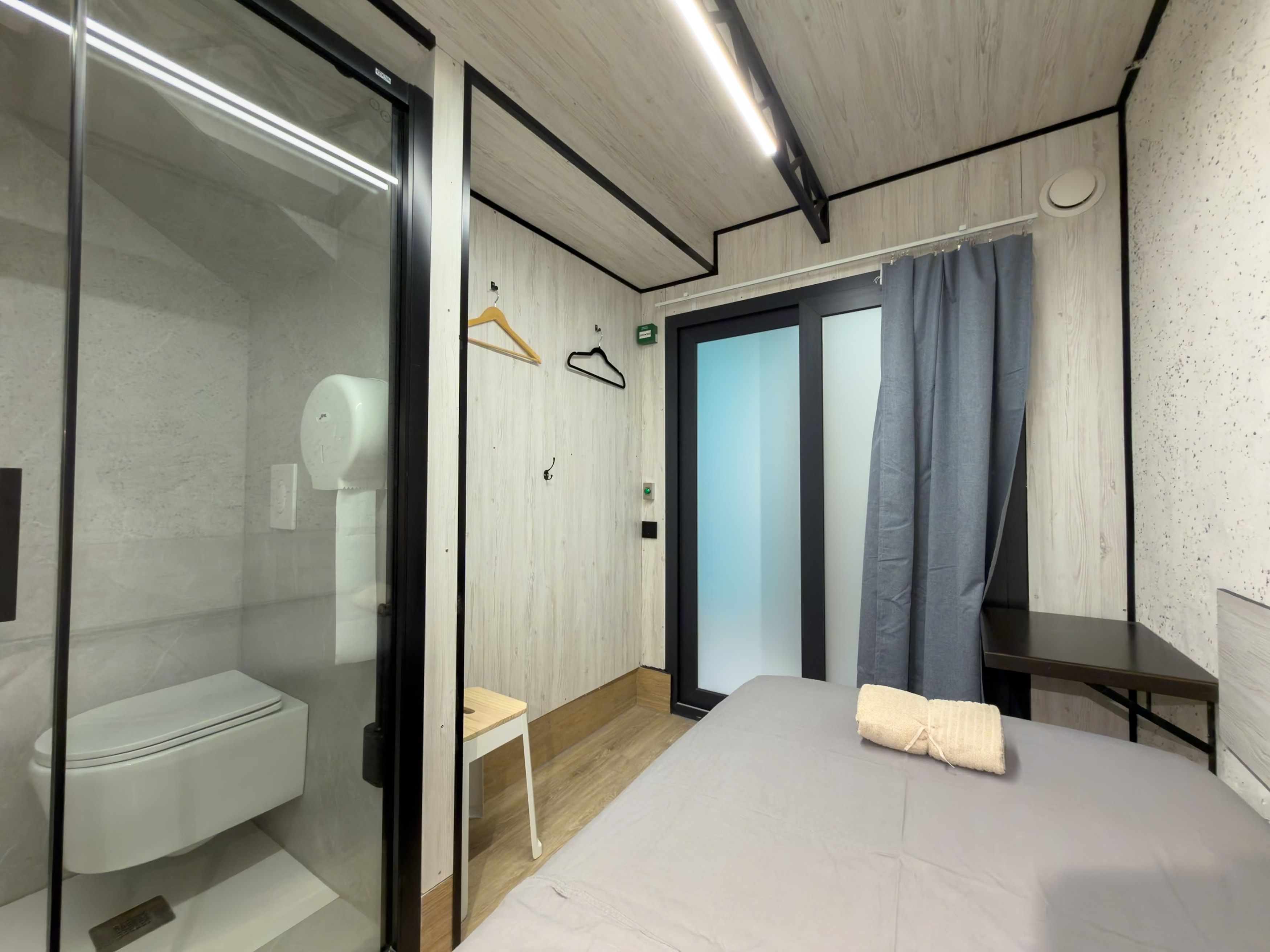 Un dormitorio moderno con cabina de ducha de vidrio, suelo de madera, cama con manta color beige y una mesita de noche pequeña.