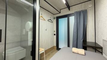 Un dormitorio moderno con cabina de ducha de vidrio, suelo de madera, cama con manta color beige y una mesita de noche pequeña.