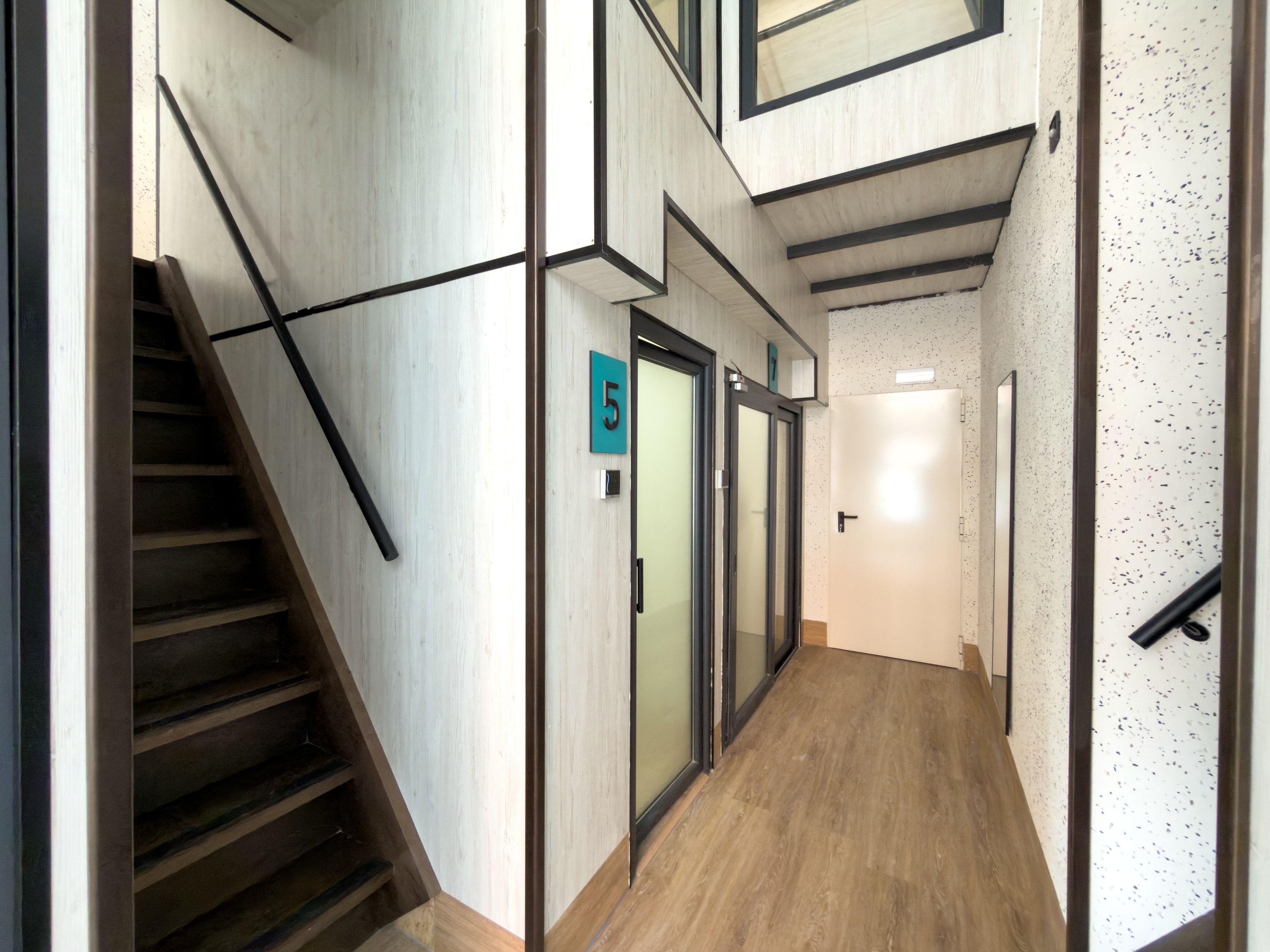 Un interior moderno con una escalera de madera, un pasillo con una puerta marcada con el número '5' y una pared con textura moteada.