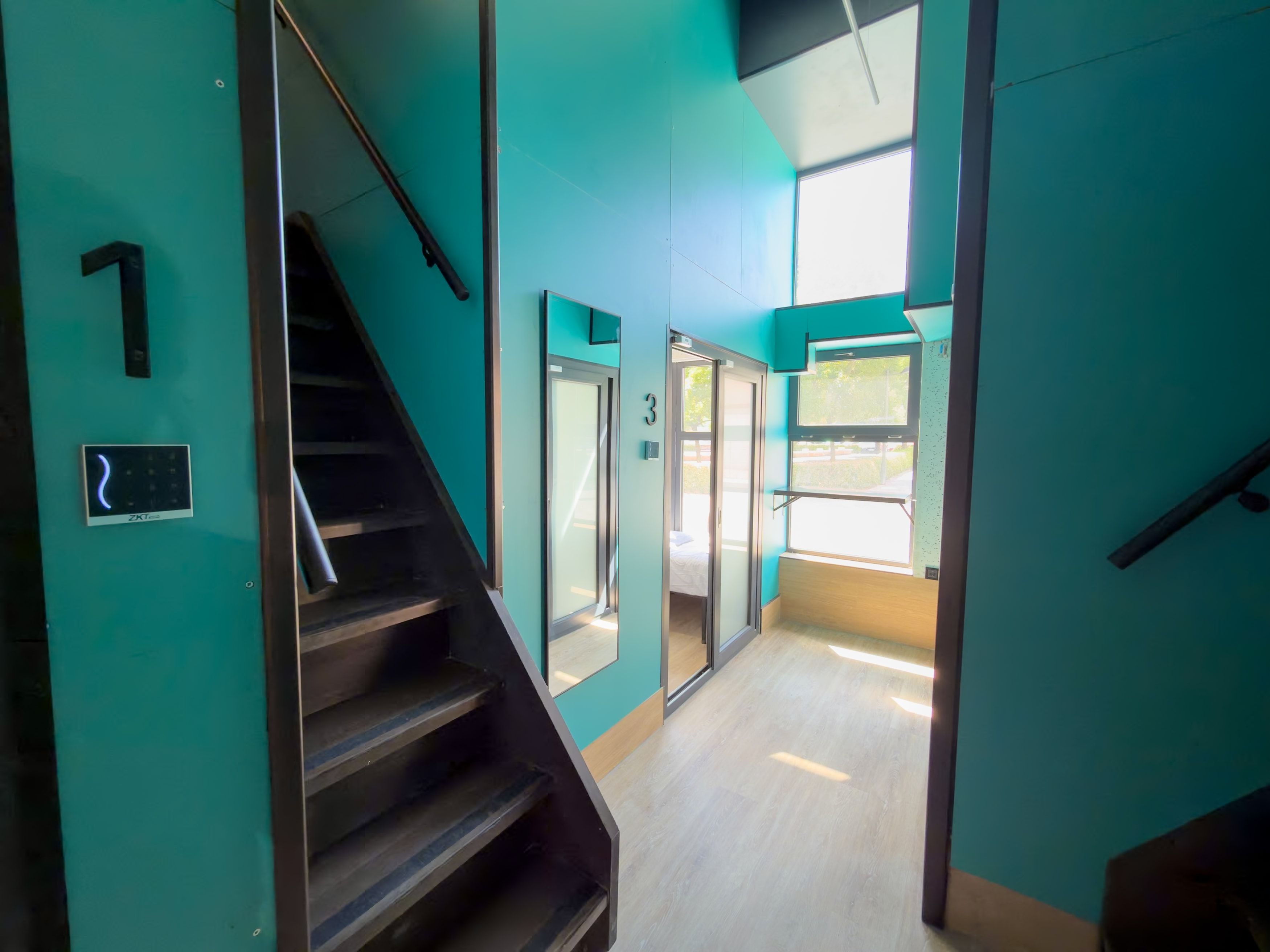 Una escalera conduce a una habitación con una cama, una puerta numerada con el número 3 y un ventanal que proporciona luz natural.
