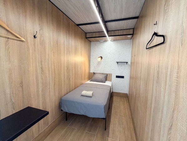 Une petite pièce comprenant un lit, un banc et une étagère fixée au mur.
