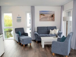 A modern living room with a grey sofa, armchairs, and a coffee table. There are large windows with curtains, a painting of a beach, and a sign on the wall.