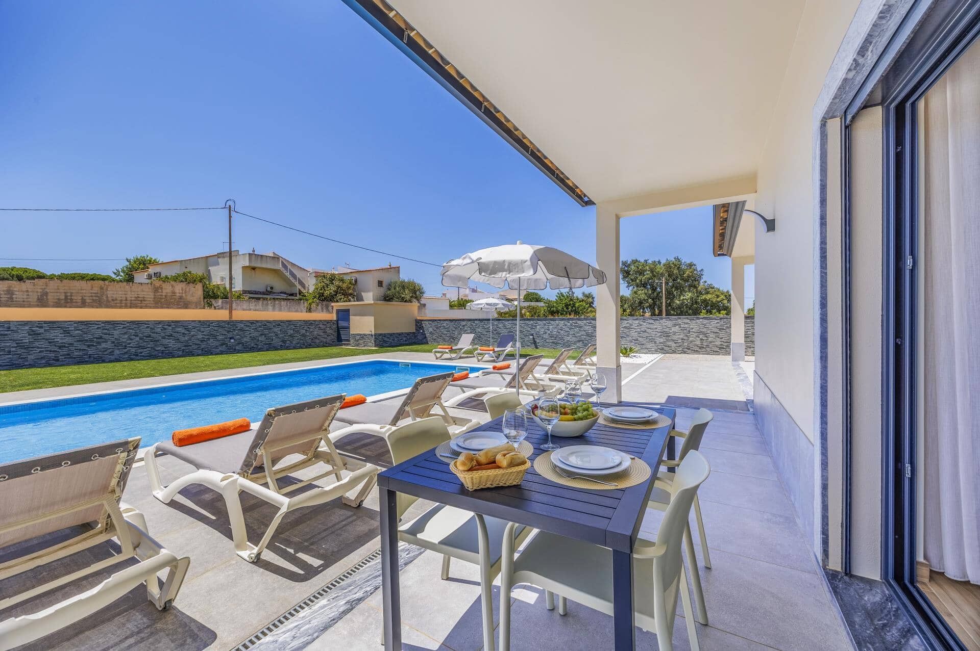 A poolside dining area with a table set for two, lounge chairs, and a view of a swimming pool.