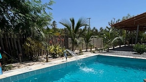 A clear blue swimming pool surrounded by a tiled deck, with palm trees and a wooden pergola in the background.