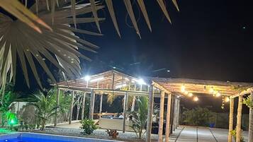 Área externa noturna com piscina, espaço de estar coberto e um caminho.