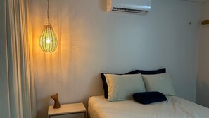 A bedroom with a bed, pillows, a nightstand, a wall-mounted air conditioner, and a hanging light fixture.