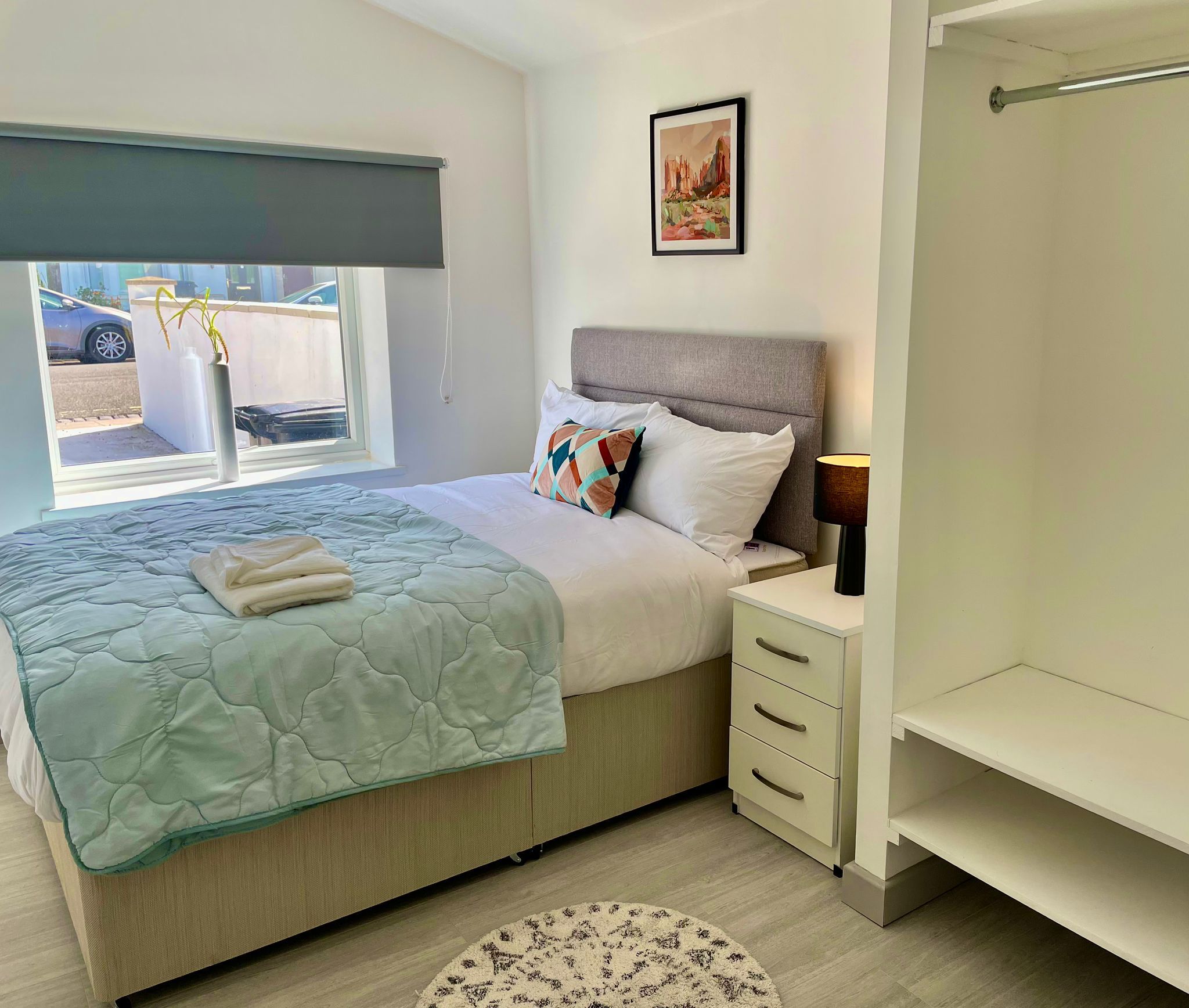 A bedroom with a bed, bedside table, lamp, and a window with a view of the outside.
