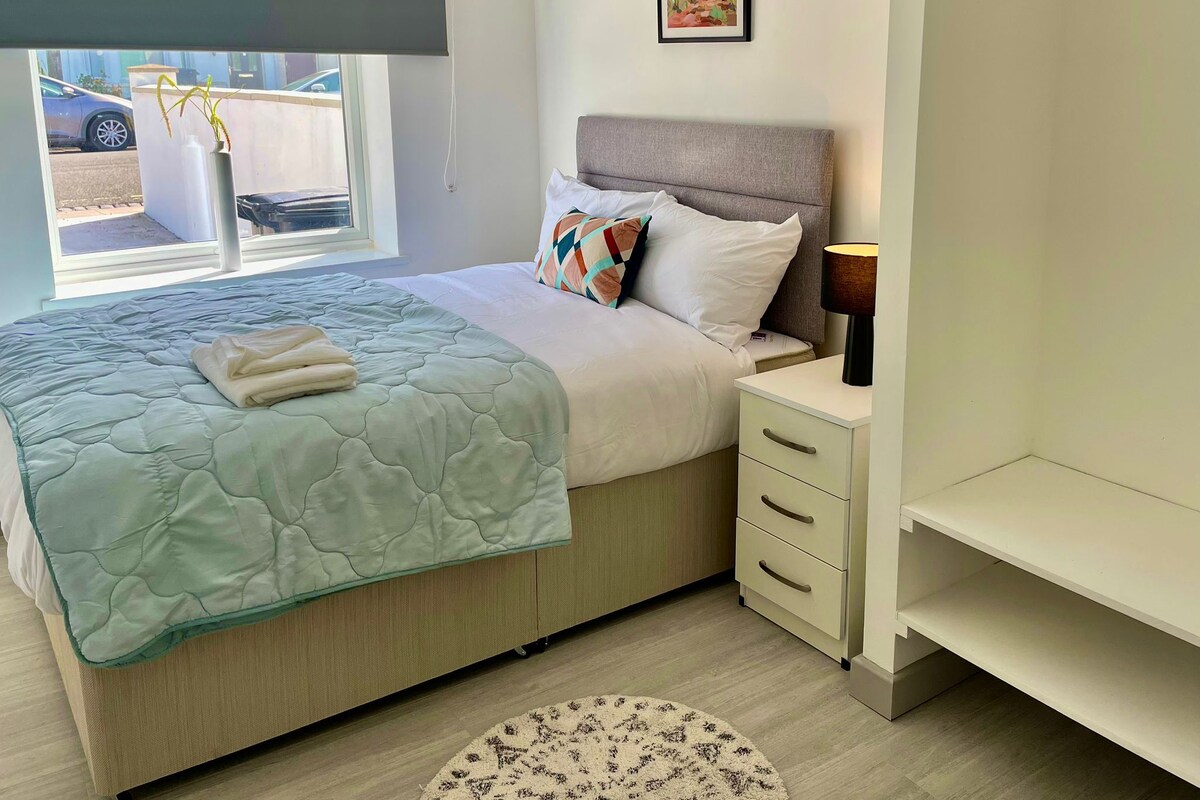 A bedroom with a bed, bedside table, lamp, and a window with a view of the outside.