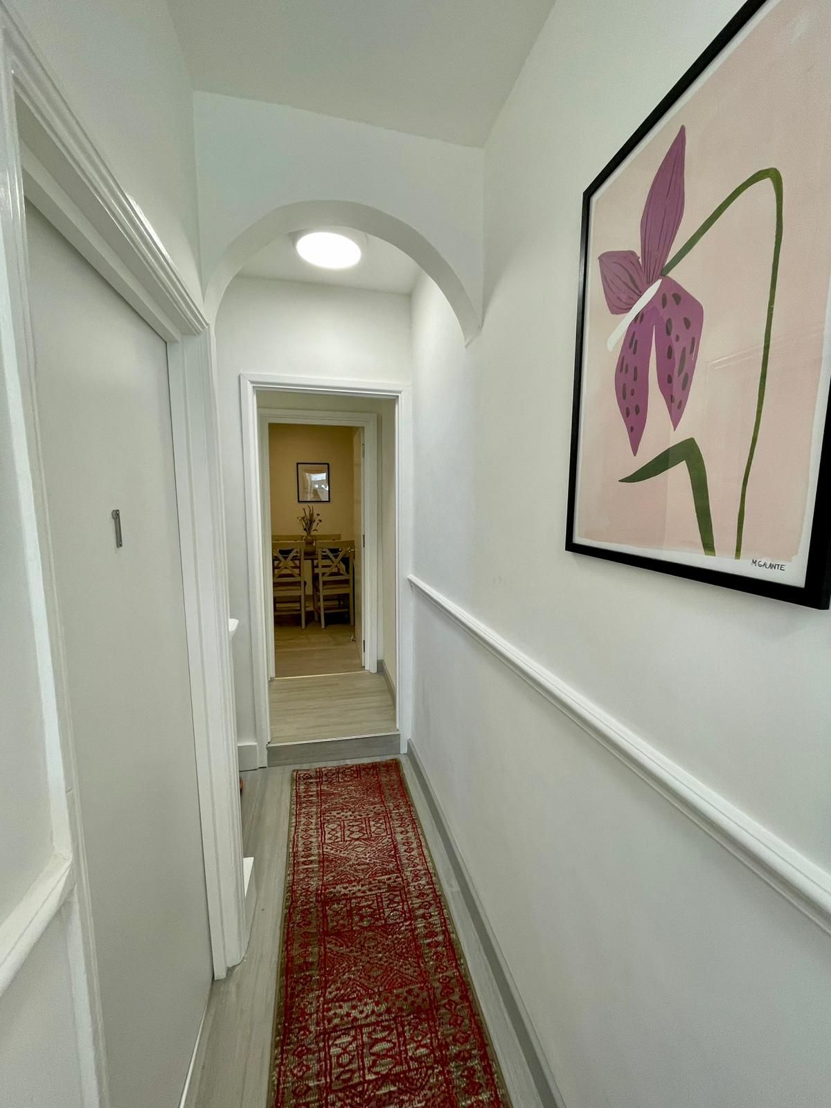 A hallway with a red patterned carpet, white walls, and a framed artwork of a purple flower.