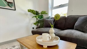 Uma sala de estar moderna com um sofá cinzento, uma mesa de centro com bandeja, uma planta em vaso e um quadro na parede.