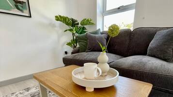 Een moderne woonkamer met een grijze bank, een bijzettafeltje met dienblad, een potplant en een lijst met foto aan de muur.