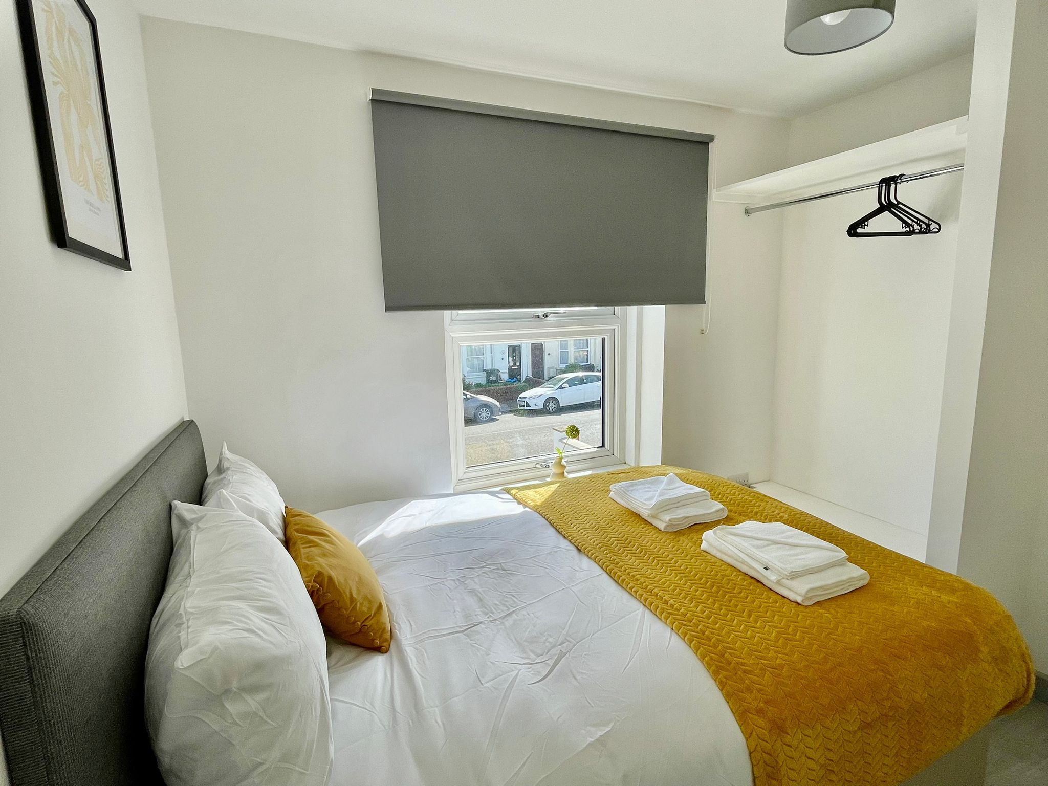 A hotel room with a bed, a window with a view of the street, a gray blind, and a hanging coat rack.