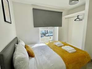 Habitación de hotel con cama, ventana que da a la calle, persiana gris y perchero.