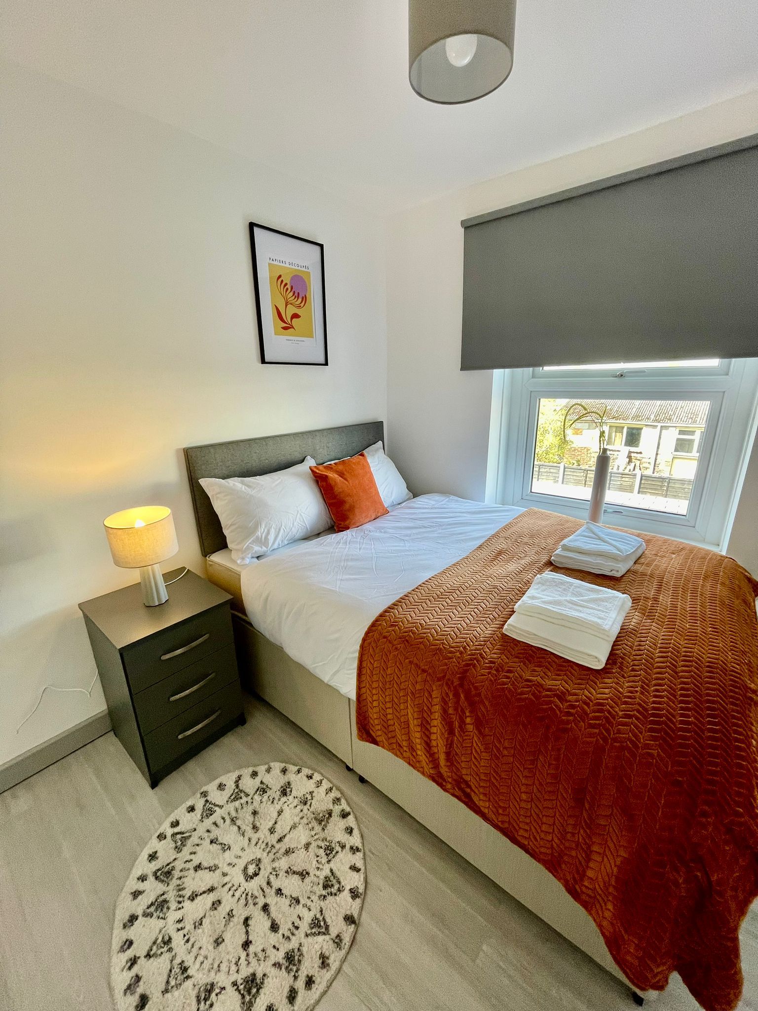 A bedroom with a bed, bedside table, lamp, artwork, window, and a view of the outside.