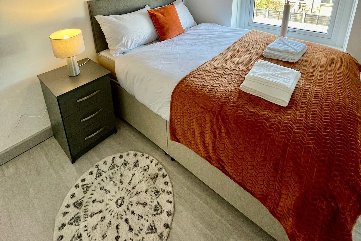 A bedroom with a bed, bedside table, lamp, artwork, window, and a view of the outside.
