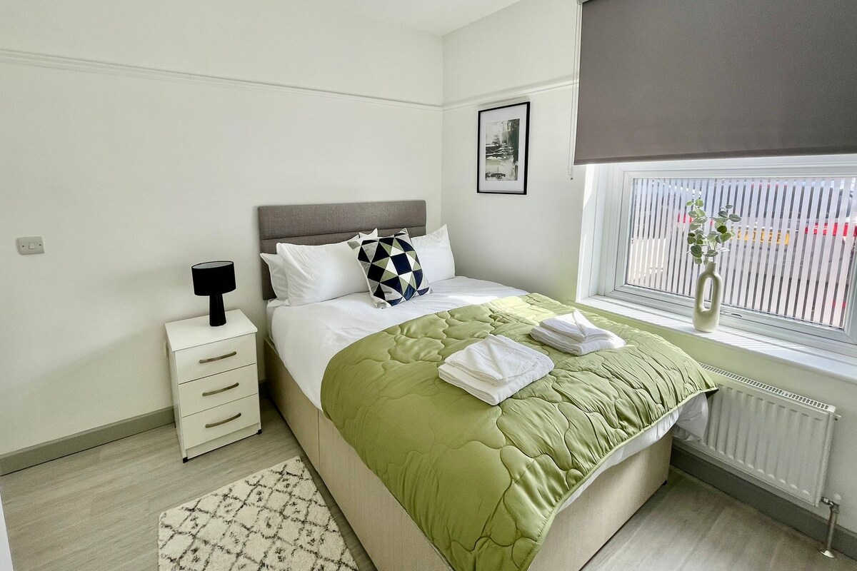 A modern bedroom with a bed, bedside table, lamp, and a window with blinds.