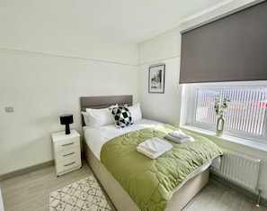 A modern bedroom with a bed, bedside table, lamp, and a window with blinds. - Ridgeways Premium House (Bristol City)