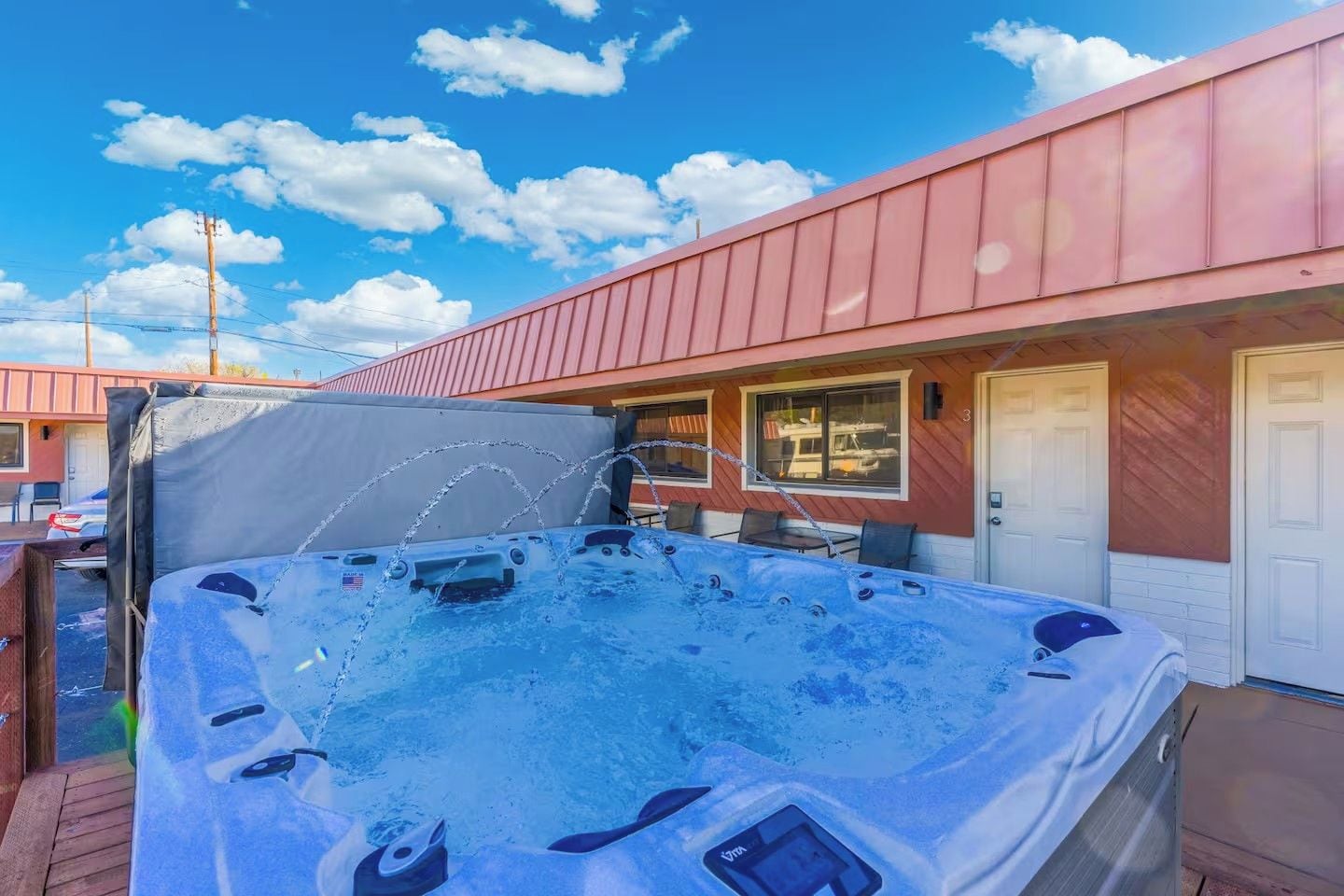 Un bain à remous sur une terrasse en bois, avec un espace abrité, un bâtiment doté d’un toit rouge et un ciel bleu clair parsemé de nuages.