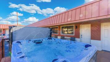 Un bain à remous sur une terrasse en bois, avec un espace abrité, un bâtiment doté d’un toit rouge et un ciel bleu clair parsemé de nuages.