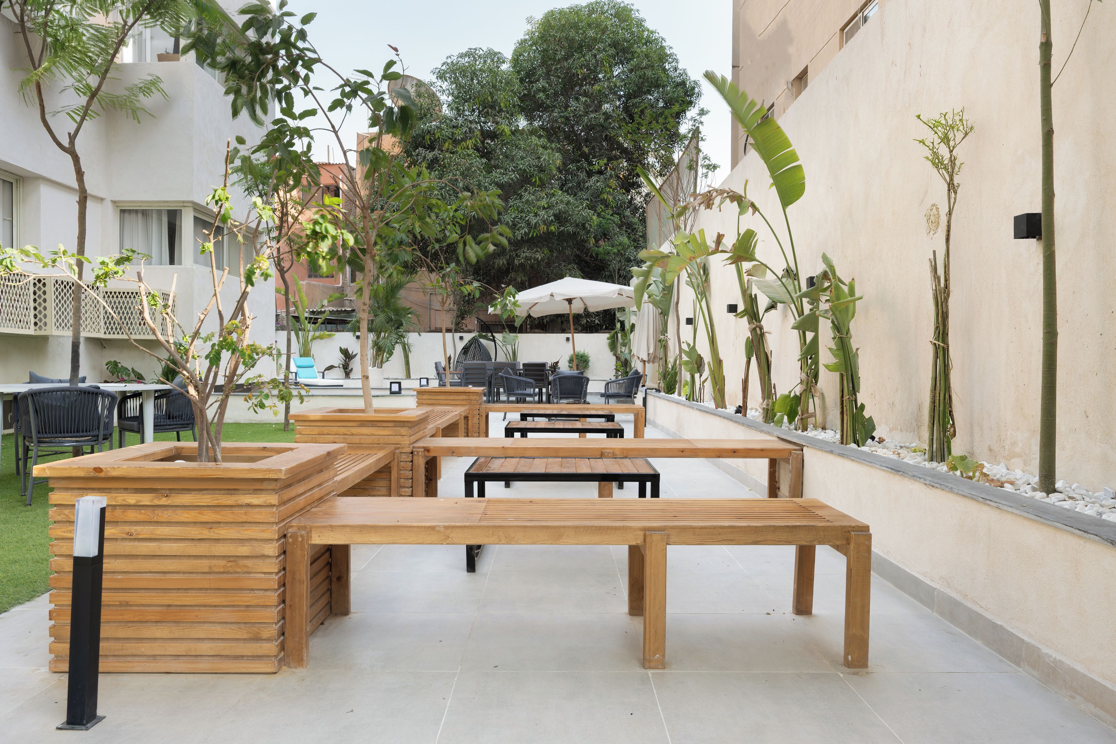 Un espace extérieur aménagé avec des tables et des chaises en bois, entouré de plantes en pot et surmonté d’un parasol blanc.