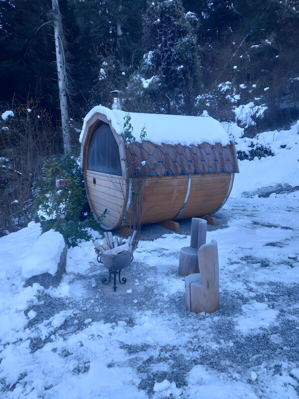 Een houten constructie bedekt met sneeuw, met een trommelvormige vorm, een kleine metalen kachel en een vuurkuil.