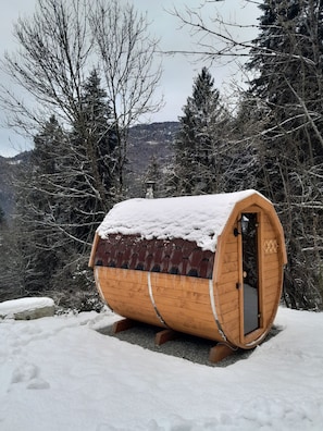 A wooden barrel-shaped structure covered in snow, with a door and a small window, surrounded by snow-covered ground and trees. - Splendid chalet with sauna and mountain views (Tione di Trento)