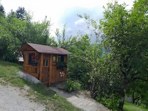 Une cabane en bois avec un toit en pente, une fenêtre et une porte, entourée de verdure et bordée d’un chemin de gravier.