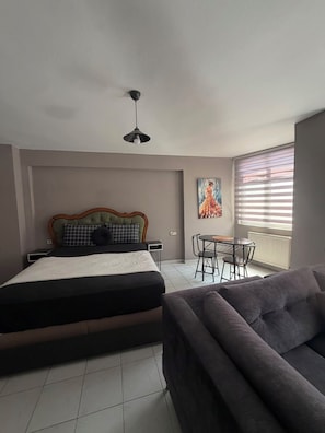 Ein Schlafzimmer mit einem Bett, einem Sofa, einem Esstisch mit Stühlen, einer Deckenleuchte und einem Fenster mit Jalousien.