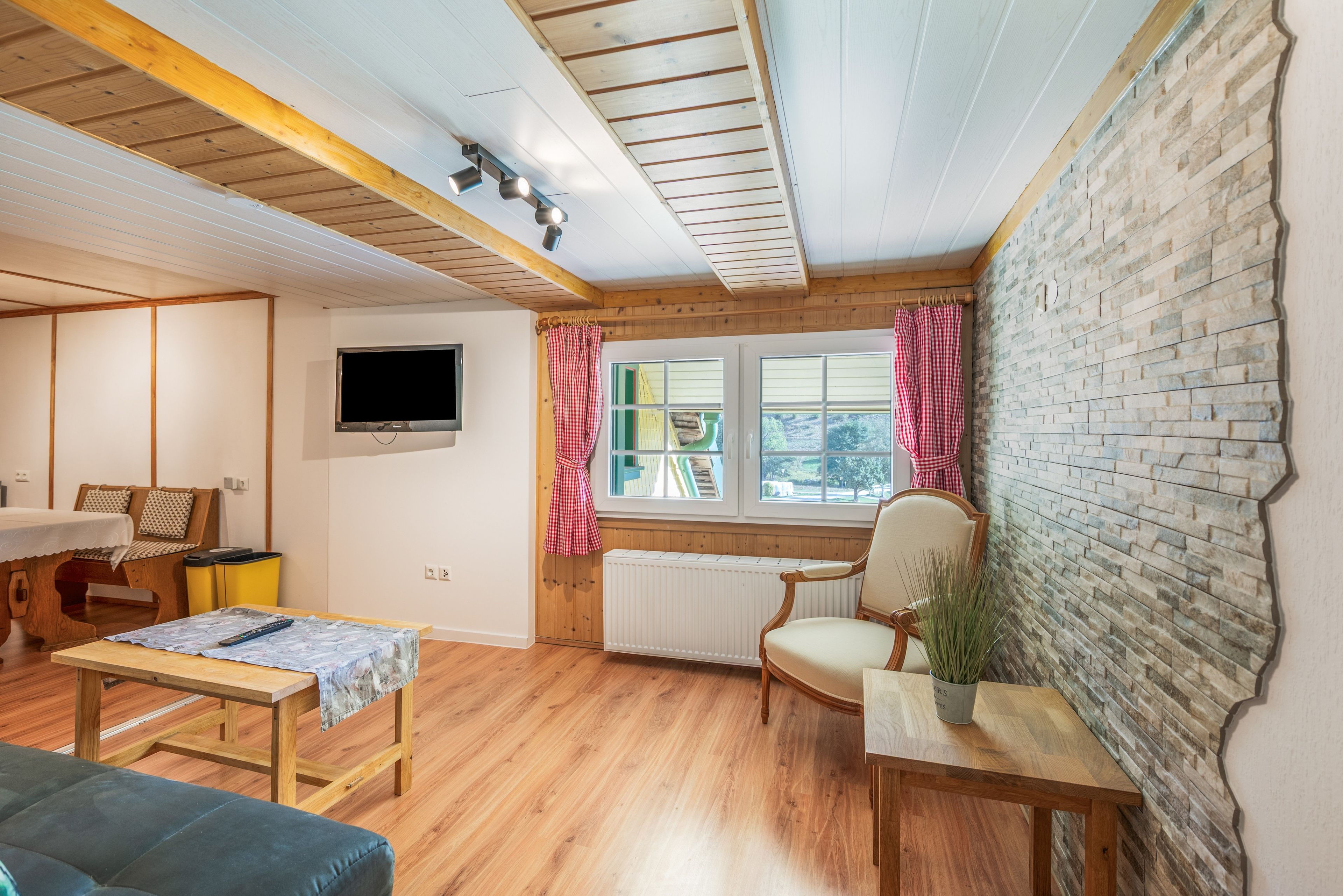 Una habitación acogedora con techo de madera, televisor instalado en la pared, ventana con cortinas, radiador y una mesita con un control remoto encima.