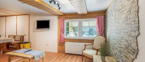 Una habitación acogedora con techo de madera, televisor instalado en la pared, ventana con cortinas, radiador y una mesita con un control remoto encima.