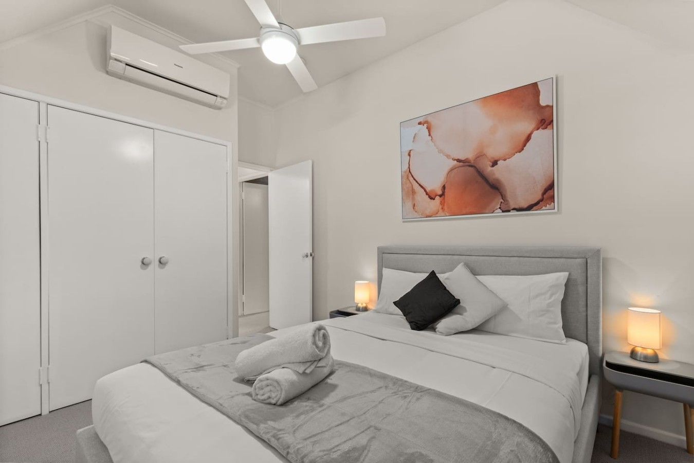 A hotel room with a bed, a ceiling fan, an air conditioning unit, a wall-mounted artwork, and bedside lamps.