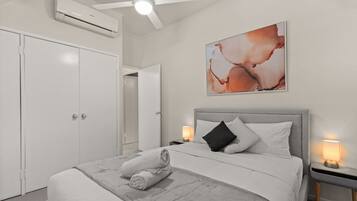 A hotel room with a bed, a ceiling fan, an air conditioning unit, a wall-mounted artwork, and bedside lamps.