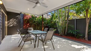 Un patio al aire libre con mesa y sillas, un ventilador de techo y vistas a un jardín con diversas plantas y una valla.