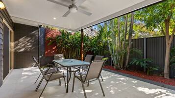 An outdoor patio with a table and chairs, a ceiling fan, and a view of a garden with various plants and a fence.