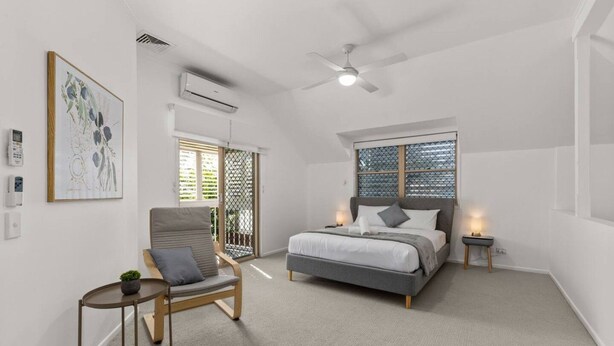 Un dormitorio con cama, una silla, una mesita de noche, ventilador de techo y ventana con persianas.