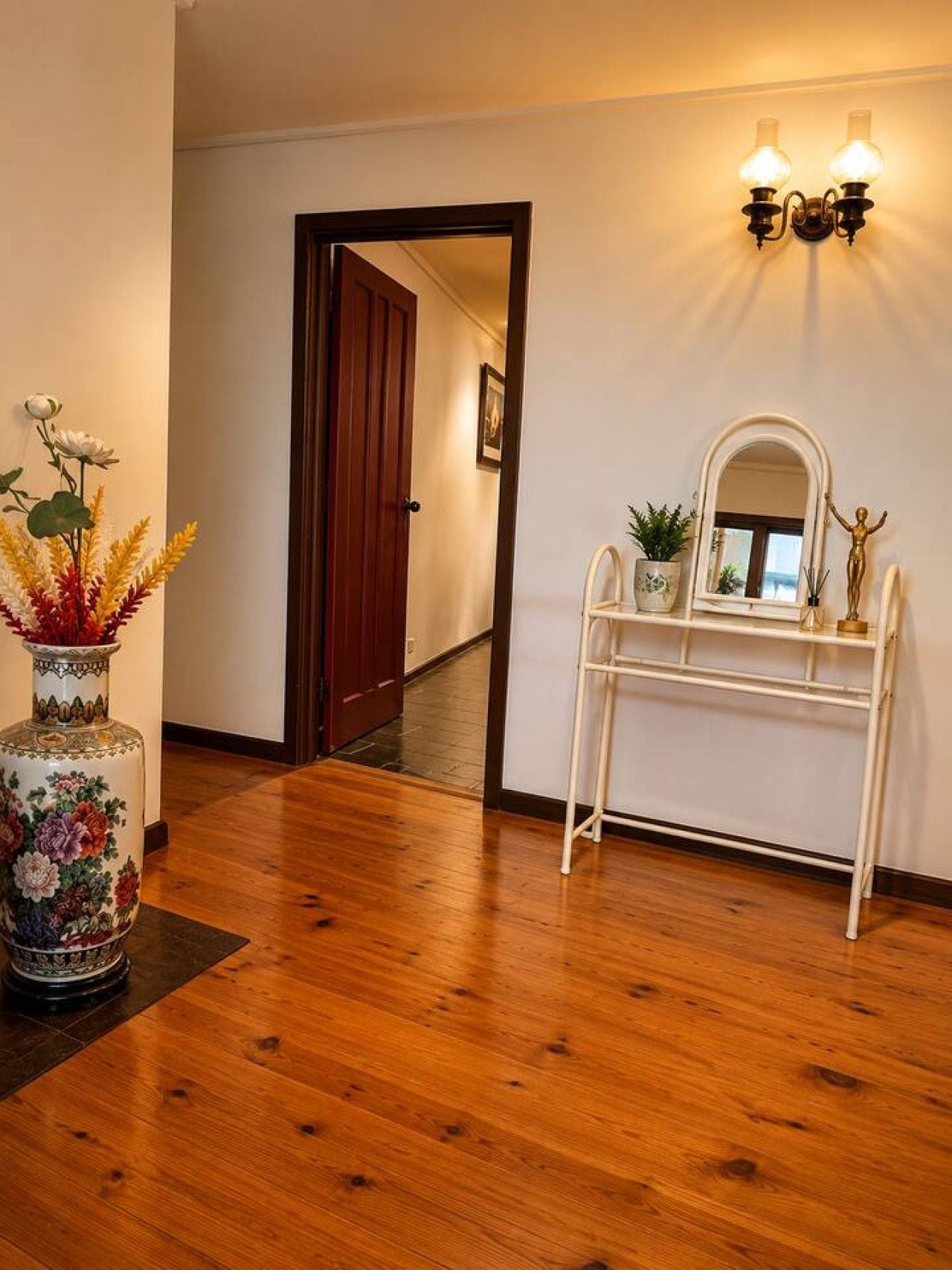 Un pasillo con piso de madera, una consola blanca con espejo y una planta pequeña, además de un jarrón decorativo con flores.