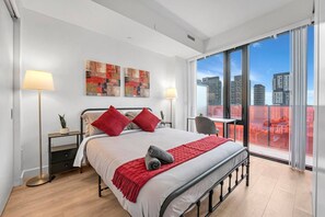 A modern hotel room with a large bed, two red pillows, a red throw blanket, a desk with a chair, and a view of the city through large windows. - *2 Bed Condo* Near UofT/Rogers/Scotia/Hospital/Gym (Toronto)